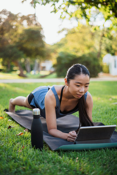 Ázsiai fitness fiatal nő csinál deszkák a parkban, miközben nézi a tutorial egy tabletta - Fotó, kép