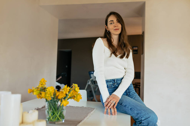 Lovely cute attractive woman with loose hair wearing white shirt and jeans is sitting in the kitchen at table with flowers in sunny day and smiling to camera - Fotó, kép