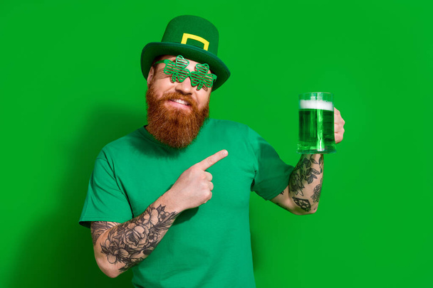 Photo of cheerful cool man wear t-shirt headwear pointing ale beer glass enjoying shamrock day isolated green color background. - Photo, image