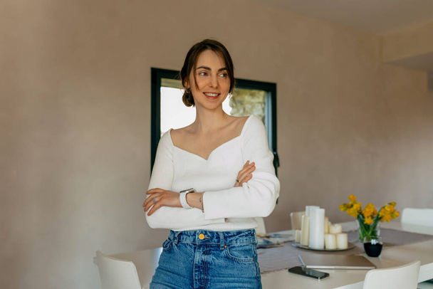 Щаслива жінка, яка носить білу сорочку і джинси, посміхається вдома. Брюнет стоїть у вітальні на задньому плані білого столу з квітами. Концепція позитивних емоцій. - Фото, зображення