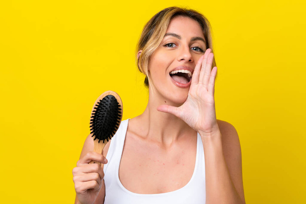 Young Uruguayan woman with hair comb isolated on yellow background shouting with mouth wide open - Foto, imagen