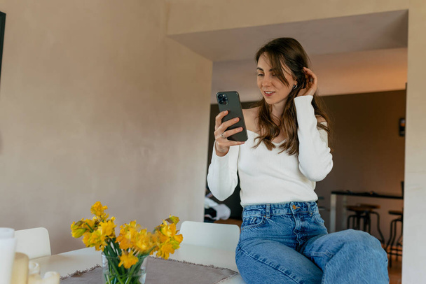 Atrakcyjna stylowa dziewczyna z długimi luźnymi włosami w białej koszuli i dżinsach używa smartfona, siedzi na stole z kwiatami i odpoczywa w domu w weekend - Zdjęcie, obraz