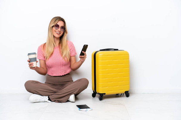 Mulher viajante uruguaia sentado no chão isolado no fundo branco segurando café para levar e um celular - Foto, Imagem