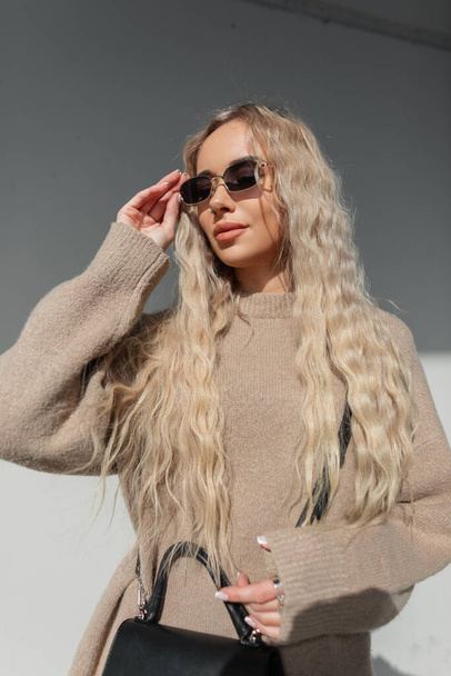 Fashionable beautiful stylish woman with a blond curly hairstyle in a fashion beige sweater wears vintage style sunglasses and walks on a sunny day on the street - Photo, Image