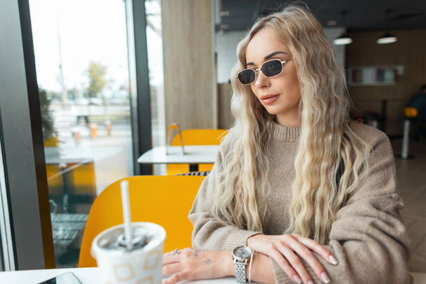 Piękna stylowa kobieta model z kręconą blond fryzurę z chłodnym vintage okulary przeciwsłoneczne w beżowym swetrze siedzi w kawiarni w pobliżu okna - Zdjęcie, obraz