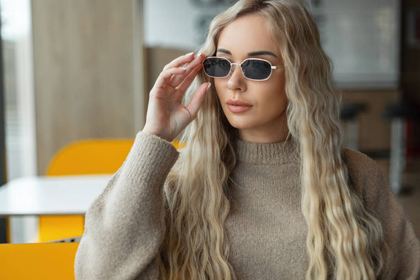 Divat gyönyörű szőke lány modell egy bézs pulóver visel vintage elegáns napszemüveg és ül egy kávézóban - Fotó, kép