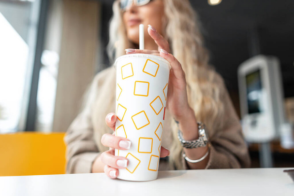 Ziemlich trendiges Mädchen sitzt in einem Café und trinkt Kaffee. Pappbecher mit Cola, Nahaufnahme - Foto, Bild