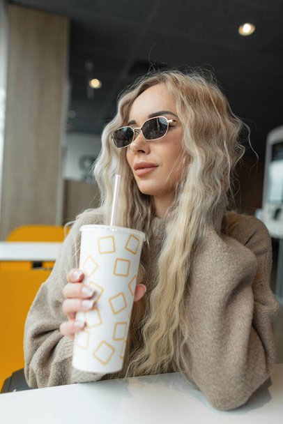 Menina bonita hipster encaracolado loira com óculos de sol vintage em roupas de moda senta-se em um café, segura uma xícara de papel de cola e bebe café - Foto, Imagem