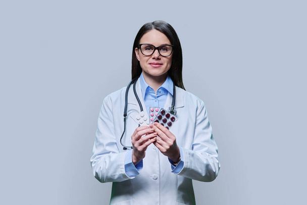 Young woman doctor recommending different pills in blister pack, on light gray studio background. Medicine, healthcare, treatment, pharmacy, pharmacology concept - Fotó, kép