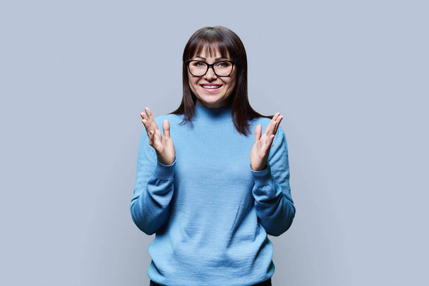 Portrait of positive mature woman with glasses, dressed in blue jumper on light gray background. Smiling 40 female looking in camera. Age, beauty, lifestyle concept - Fotó, kép