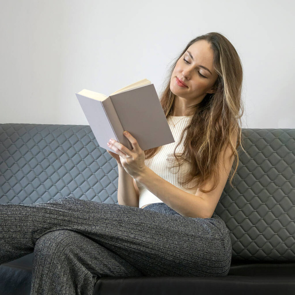 Νεαρή γυναίκα στον καναπέ χαμογελώντας και κρατώντας ένα βιβλίο, ανάγνωση, έννοιες του σπιτιού και της άνεσης, χώρος για κείμενο. - Φωτογραφία, εικόνα