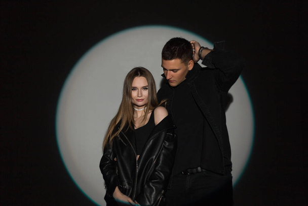 Красивая молодая пара мужчина и женщина в стильной джинсовой повседневной черной одежде с кожаной курткой и черной джинсовой рубашкой на темном фоне в светлом круге - Фото, изображение