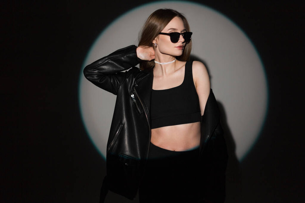 Μοντέρνο όμορφο γυναικείο hipster μοντέλο με δροσερά μαύρα γυαλιά ηλίου σε κομψά μαύρα rock ρούχα με δερμάτινο μπουφάν και top posing in σε σκούρο φόντο με κύκλο φως στο στούντιο. Όμορφο κορίτσι. - Φωτογραφία, εικόνα