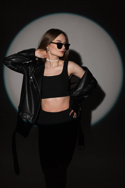 Modieuze hipster coole vrouw met trendy zonnebril in zwarte stijlvolle rock outfit met leren jas en top op een donkere achtergrond met cirkel licht op partij - Foto, afbeelding