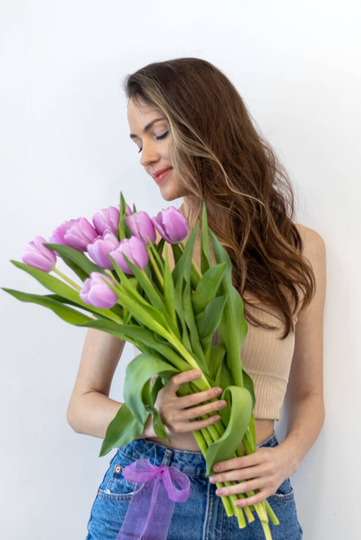 Красивая счастливая женщина с лиловыми тюльпанами стоит на белом фоне. Девушка смотрит на цветы. - Фото, изображение
