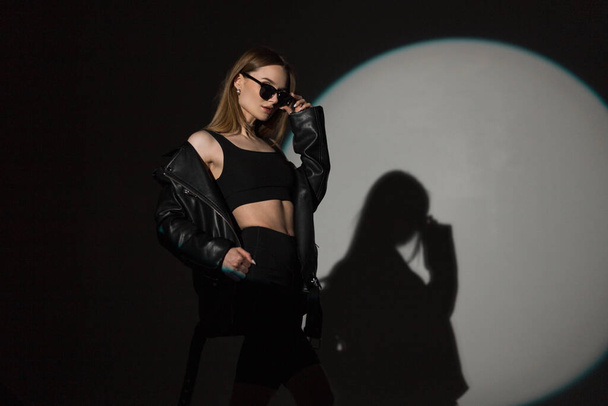 Cool elegáns csinos nő hipster divatos divat rock ruhák felső és bőr kabát visel divatos napszemüveg és pózol a sötét háttér körkörös fény és árnyékok a stúdióban - Fotó, kép