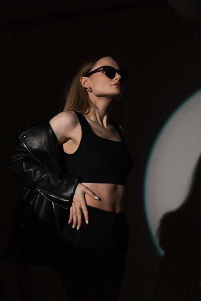 Cool divat hipster gyönyörű nő napszemüveg elegáns fekete felső és divatos bőrkabát pózol a sötétben a stúdióban - Fotó, kép