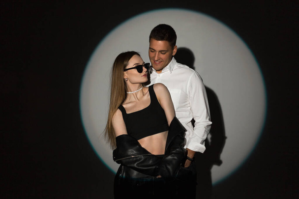 Stylish pretty young couple beautiful girl and handsome man in fashion clothes with leather jeans and white shirt on a dark background with a circle of light at a party - Foto, Imagen