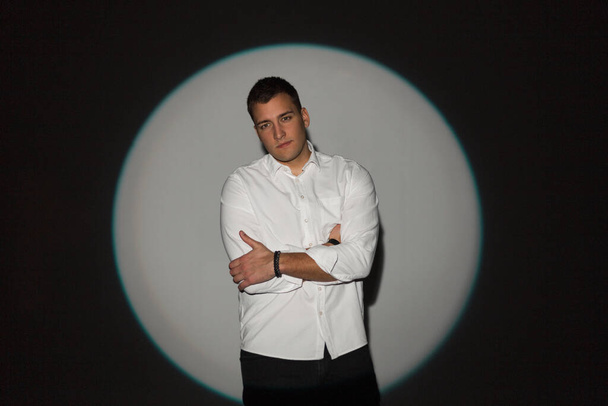 Stylischer junger erfolgreicher gutaussehender Geschäftsmann im modischen weißen Hemd steht im Atelier auf einem runden Licht vor dunklem Hintergrund - Foto, Bild