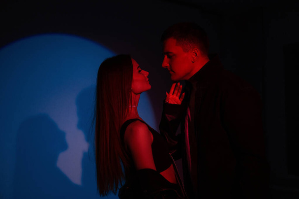 Moda bella coppia uomo e bella donna in nero abiti eleganti su uno sfondo scuro con luce rossa e blu creativa in studio - Foto, immagini