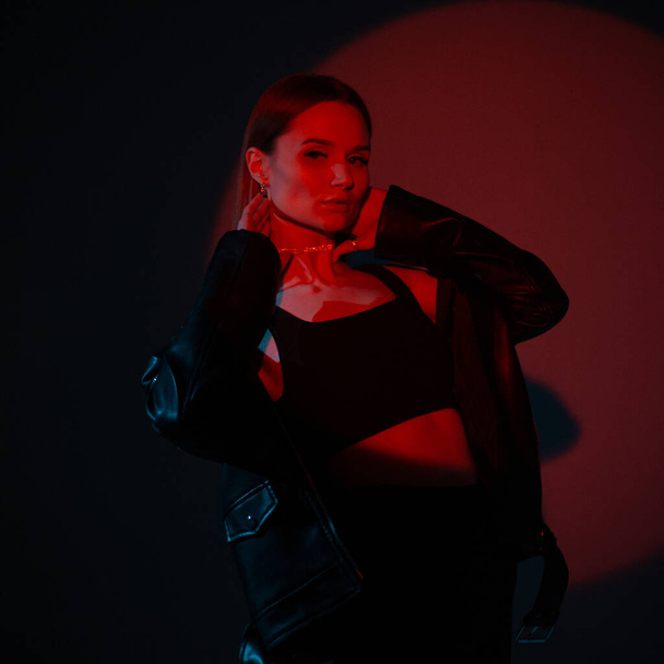 Élégante belle jeune femme à la mode vêtements noirs posant sur un fond sombre dans un studio avec lumière rouge créative - Photo, image