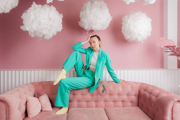 Een mooi meisje in een business pak van malachiet kleur zit op een bank of bank in een pastel roze kamer met zwevende wolken. - Foto, afbeelding