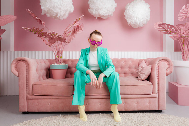 Een mooi meisje in een business pak van malachiet kleur zit op een bank of bank in een pastel roze kamer met zwevende wolken en bedekt met roze indoor plant. - Foto, afbeelding
