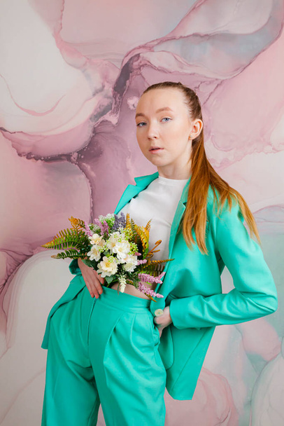 A beautiful girl in a business suit of malachite color posing with a plant or flowers againt a creative pink background. - Foto, afbeelding