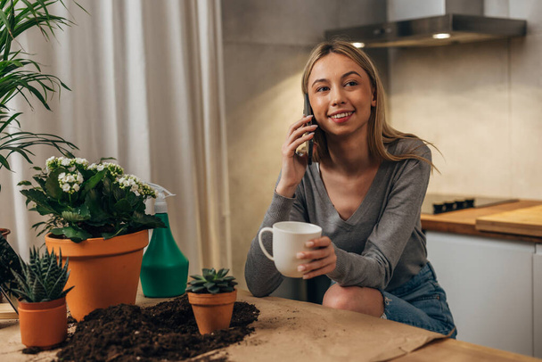 A pretty blond woman is enjoying a phone call after planting her houseplants - Фото, изображение