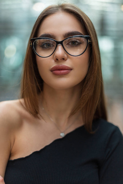 Stylowy portret pięknej, odnoszącej sukcesy młodej menadżerki w modnych okularach w modnej czarnej bluzce na jednym ramieniu w biurowym szklanym budynku - Zdjęcie, obraz