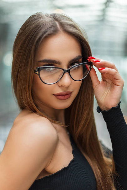 Beautiful professional business manager woman in a black elegant top puts on fashion glasses in a glass office building - Фото, изображение