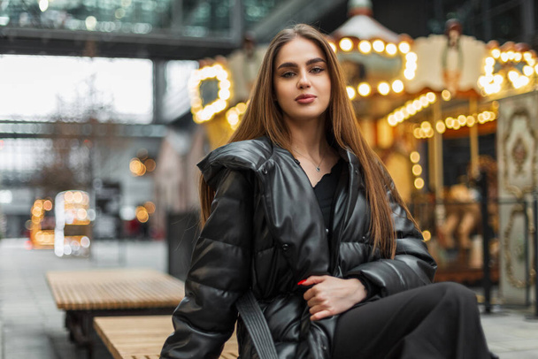 Happy beautiful young woman in fashion elegant black clothing with a down jacket sits on a bench outside near a vintage carousel with lights - Foto, Imagem