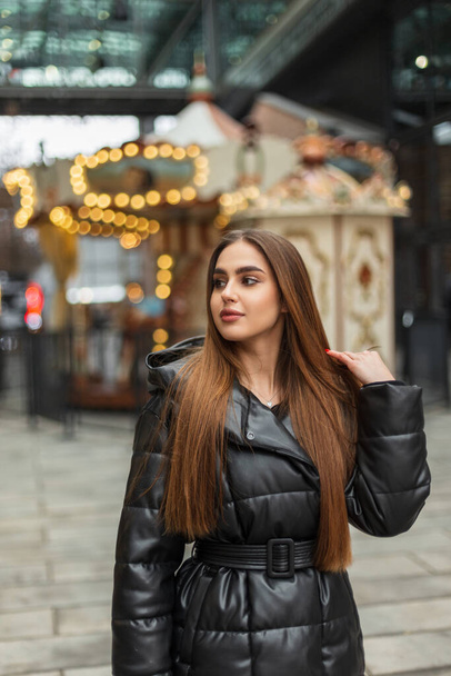 Beautiful happy young brunette woman in a black fashionable down jacket walks in the city on the background of a vintage carousel with lights - Fotó, kép