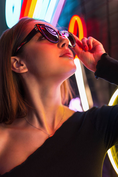 Fashion female portrait of a beautiful stylish woman near a neon light wearing fashionable black sunglasses - Fotó, kép