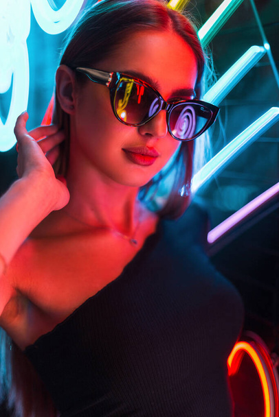 Mladá krásná módní žena se stylovými slunečními brýlemi v černých šatech na tmavém pozadí s jasnými barevnými neonovými světly. Městská dívka krása - Fotografie, Obrázek