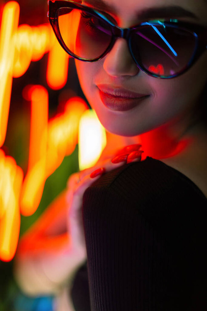 Night female portrait of a beautiful stylish girl with sunglasses on a dark background with colorful neon lights - 写真・画像