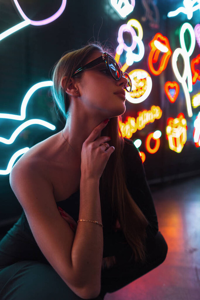 Mode belle jeune femme modèle avec des lunettes de soleil en noir vêtements élégants assis et pose sur un fond sombre avec des néons - Photo, image