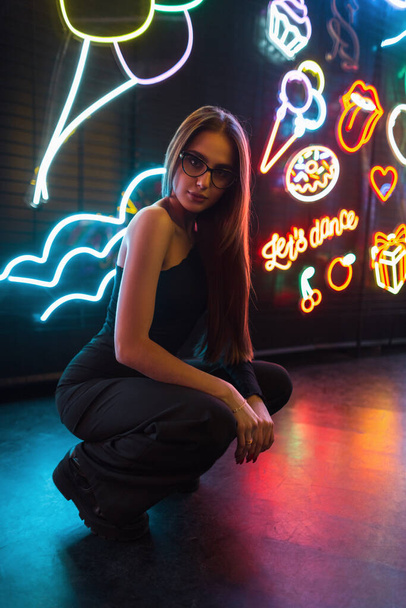 Модная красивая модель в очках и черной модной одежде сидит на темном фоне с яркими разноцветными неоновыми вывесками - Фото, изображение