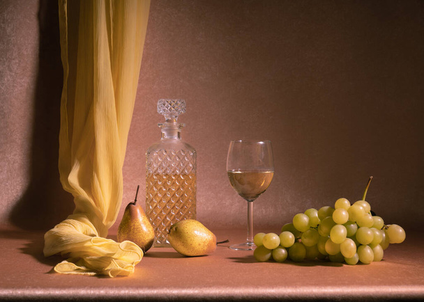 Eine Karaffe Weißwein neben einem Glas Weißwein, zwei Birnen und einem Bund weißer Trauben auf orangefarbenem Hintergrund. Mit einem gelben Schal. Stillleben. - Foto, Bild