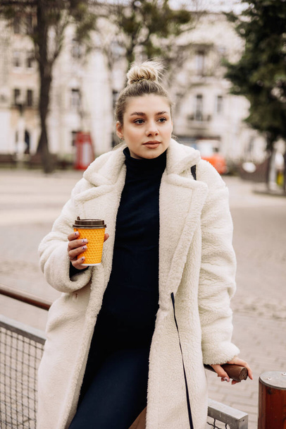 A cute young woman in a white fur coat and a cup of coffee walks through the city. Stylish young pregnant girl. Women's casual fashion - Photo, image