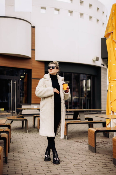 A young pretty woman in a white fur coat and sunglasses with coffee walks around the city. Stylish young girl. - Photo, Image