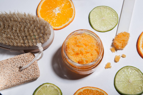 An orange scrub in a glass jar on a white table with pieces of citrus fruits. Nearby is a brush made of natural bristles for body peeling, a pumice stone for feet. - Photo, Image