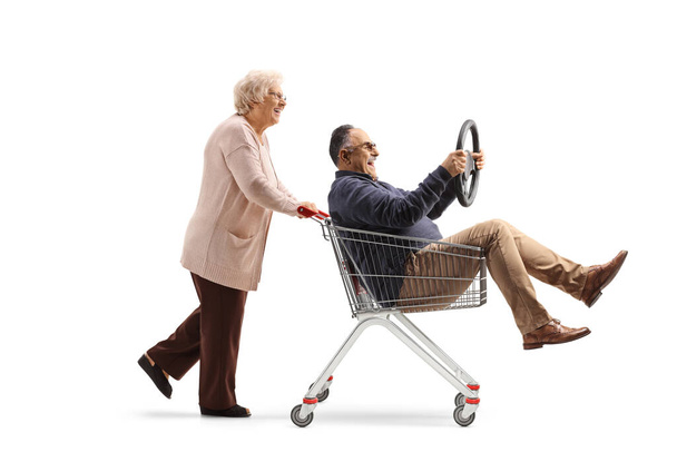Mujer mayor empujando a un hombre maduro dentro de un carrito de compras sosteniendo un volante aislado sobre fondo blanco - Foto, Imagen