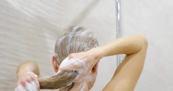 Jeune femme sous un jet chaud de douche, elle masse les cheveux de sa tête avec ses mains. Se laver la tête avec un shampooing. Soins pour les beaux cheveux longs. - Séquence, vidéo