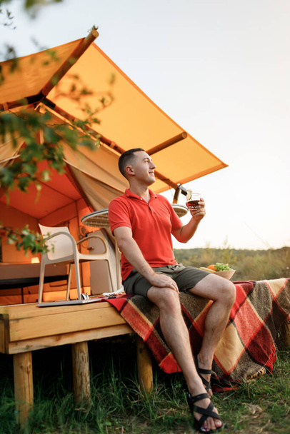 Happy man relaxing in glamping on summer evening, drinking wiskiey with cola. Luxury camping tent for outdoor recreation and recreation. Lifestyle concept - 写真・画像