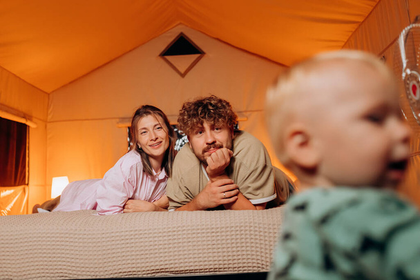 Happy family with lovely baby playing and spend time together in cozy glamping on summer evening while lying on bed. Luxury camping tent for outdoor recreation and recreation - Zdjęcie, obraz