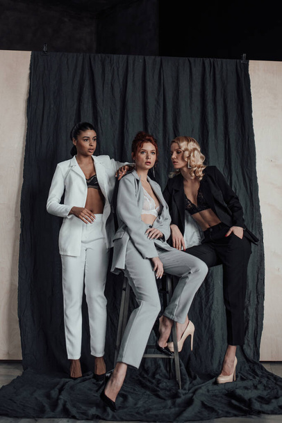 Adult beauty elegant multi ethnic group young girls in formal evening suits of different colors with bra. Stylish diverse people sensual fashionistas posing at studio in fashion pantsuits no shirts - Fotografie, Obrázek