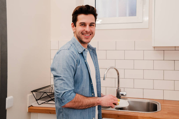 若い白人男性が台所で皿を洗う。家事掃除をしているハンサムな笑顔の男とカメラを見て..ハウスキーピングの概念。高品質の写真 - 写真・画像