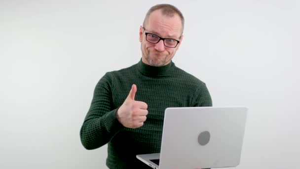 adult man in glasses with a beard and a mustache is very happy he shows a thumbs up in his hands he has a gray laptop on a white background 50-60 years old green sweater good luck success victory - Materiał filmowy, wideo