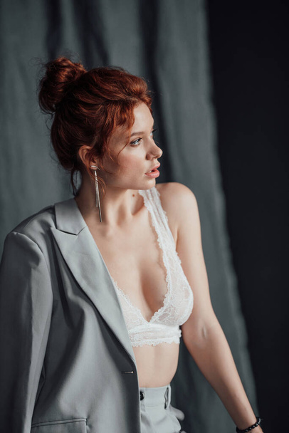 Adult beauty elegant young woman in formal evening gray suit with white bra at thoughtful. Stylish ginger curly hair sensual model bare shoulder fashionista posing at studio in fashion pantsuit - 写真・画像